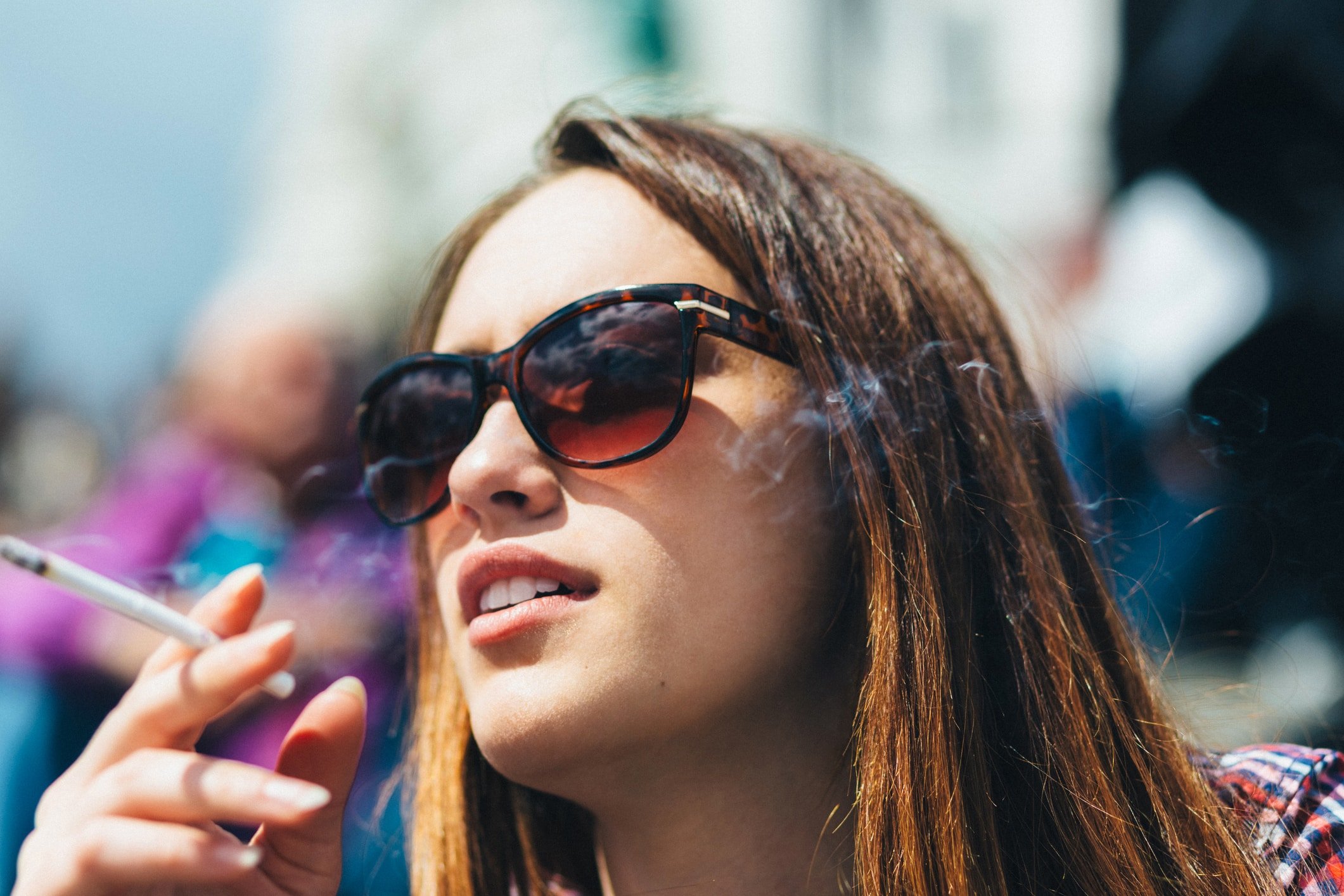 Close-up of a young woman wearing sunglasses and smoking a cigarette on a sunny day.