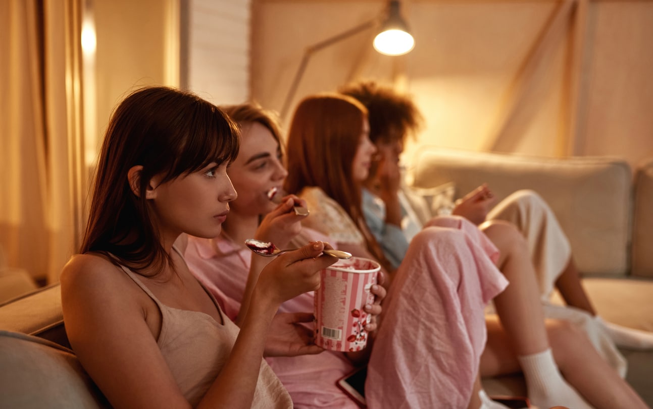 Young multiethnic girlfriends eating ice cream and watching TV or movie on sofa during girlish sleepover at home. Black and european girls. Friendship. Rest, entertainment and leisure. Yough lifestyle