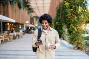 Happy young man walking outdoors checking phone while holding a coffee cup. Concept of urban lifestyle, technology, and communication